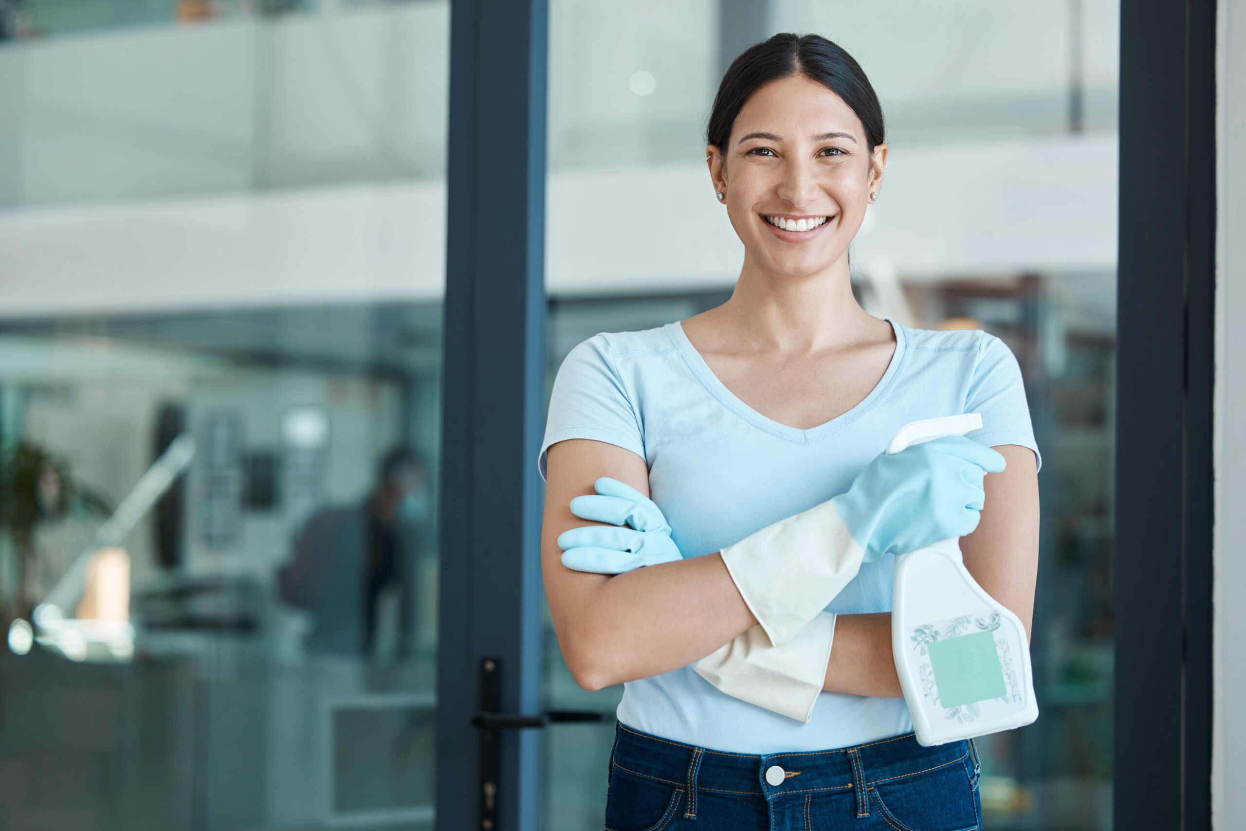 Cleaning service, portrait and cleaner in an office with spray bottle of disinfectant, bleach or detergent. Happy, smile and young female worker with gloves and soap liquid done washing glass windows.